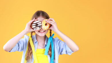 Girl in pigtails standing in front of a yellow background smiling and holding donuts in front of her eyes