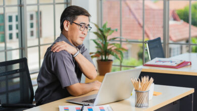 Man sits at desk with laptop rubbing shoulder.